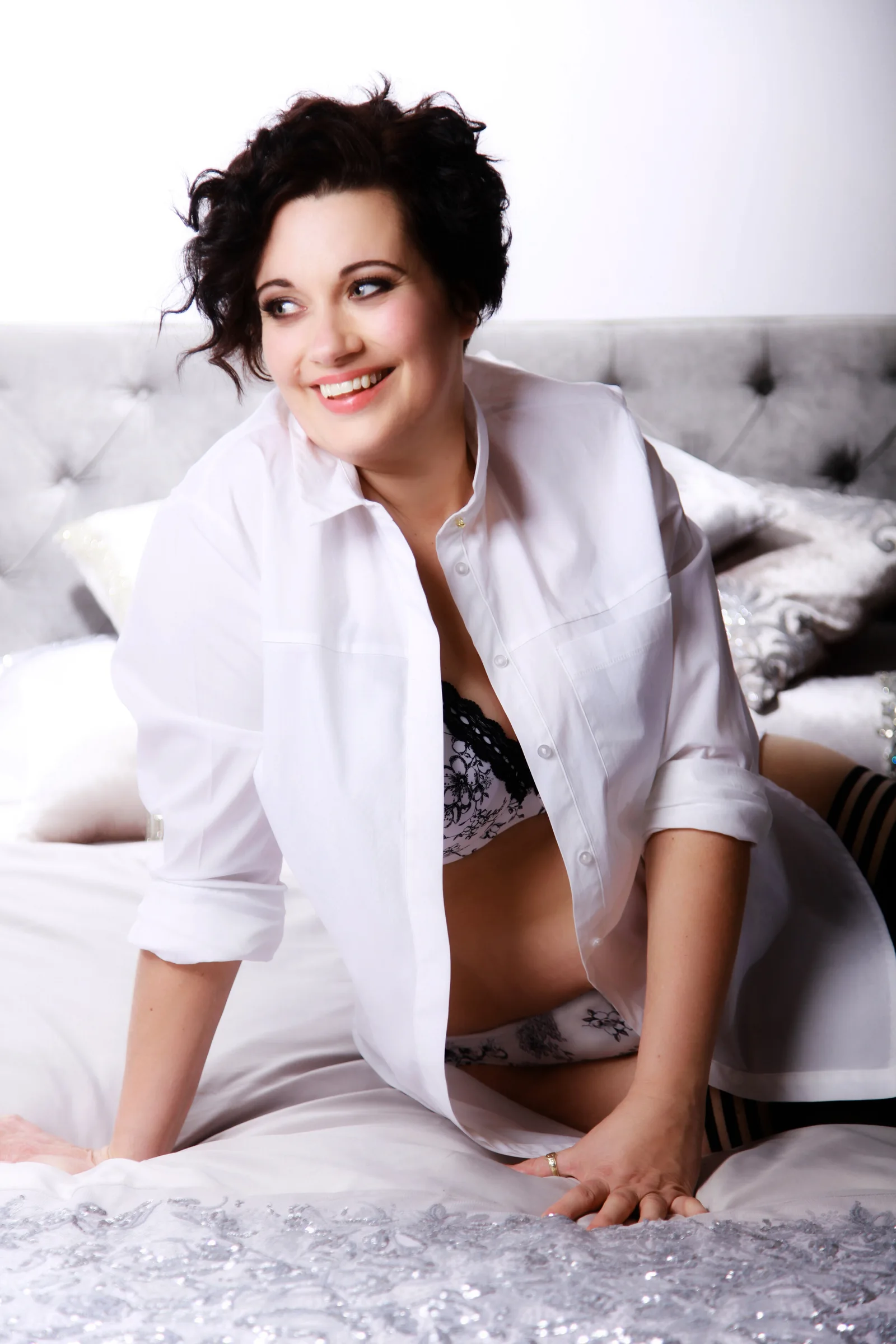 A client smiling in an open white shirt as she kneels on a bed