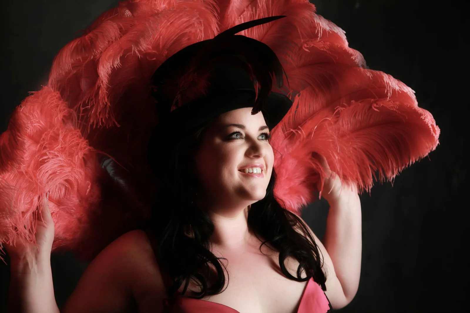 A smiling woman wearing a black top hat against a fan of red feathers